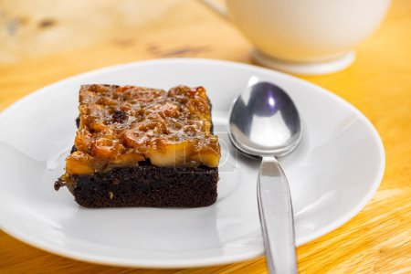 Photo for High angle view of a piece of delicious homemade toffee cake and metal spoon in white ceramic dish with a cup of coffee on wooden table. - Royalty Free Image