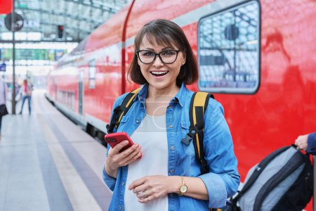 Photo for Woman waiting for railway public electric transport on platform of city station. Female with smartphone using banking app to buy online ticket payment, online timetable and route service, technology - Royalty Free Image