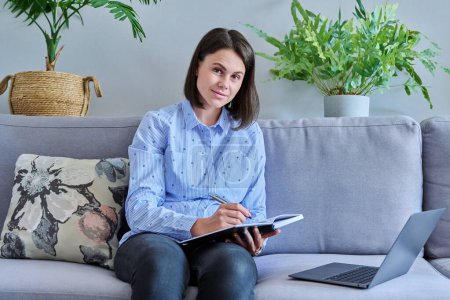 Photo for Young business woman working from home using laptop making notes looking at camera sitting on sofa in living room. Freelance remote work online business technology sales e-learning e-education concept - Royalty Free Image