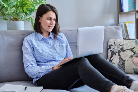 Photo for Young business woman working from home using laptop sitting on sofa at living room. Freelance, remote work, online business, technology, sales, e-learning, e-education concept - Royalty Free Image