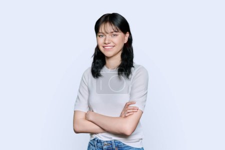 Portrait of confident positive young teenage female student with arms crossed over white studio background. Beautiful brunette woman in white clothes looking at camera with toothy smile