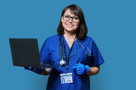 Freundliche Ärztin mit Laptop, die in die Kamera auf blauem Studiohintergrund blickt. Lächelnde Frau in blauer Uniform mit Stethoskop, Ausweis, der im Studio posiert. Gesundheitswesen, Medizin, Personalkonzept