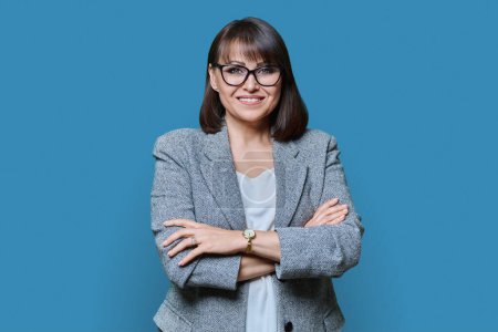 Photo for Business positive middle age woman with arms crossed on blue studio background. Confident woman with toothy smile in glasses jacket looking at camera. Business management leadership staff 40s people - Royalty Free Image