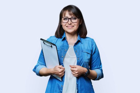 Middle aged business woman with clipboard looking at camera on white studio background. Mature smiling female teacher agent auditor psychologist supervisor posing with papers documents contracts