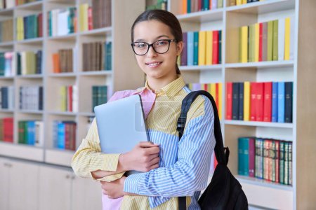 Photo for Portrait of teenager student girl looking at camera in library. Smiling teenage female 15, 16 years old wearing glasses, holding laptop backpack. High school, education, knowledge, adolescence concept - Royalty Free Image
