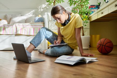 Teenage girl with glasses studying at home using a laptop. Female with headphones and textbooks sitting on the floor in the room. Education, adolescence, high school concept