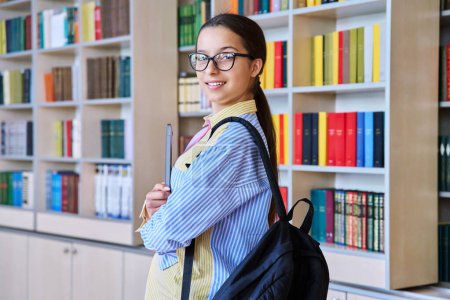 Photo for Portrait of teenager student girl looking at camera in library. Smiling teenage female 15, 16 years old wearing glasses, holding laptop backpack. High school, education, knowledge, adolescence concept - Royalty Free Image