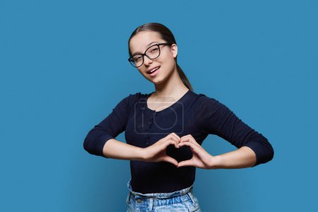 Young smiling female showing heart gesture with fingers, happy female showing love, blue studio background. Body language, signs symbols, love, emotions, romance youth concept