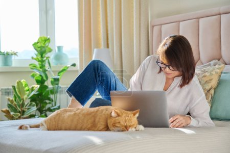 Téléchargez les photos : Femme mature utilisant un ordinateur portable assis sur le canapé à la maison, avec chat roux endormi détendu. Femme d'âge moyen travaillant à domicile à distance, pigiste, technologies pour le travail et les loisirs, 40 ans, mode de vie - en image libre de droit
