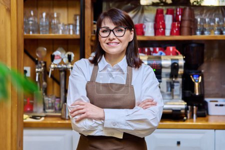 Téléchargez les photos : Portrait of middle aged woman bistro restaurant manager owner in an apron, confident smiling looking at camera with crossed arms. Small business, service, work, staff concept - en image libre de droit