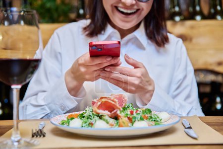 Foto de Happy woman in a restaurant taking pictures of food with a smartphone. Lifestyle, photo for social networks, personal blog concept - Imagen libre de derechos