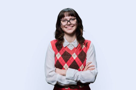 Portrait of confident pretty female student with arms crossed against light studio background. Smiling attractive cute girl in glasses 18, 19 years old looking at camera