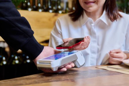 Photo for Woman in restaurant sitting at table with food paying for order using smartphone and wireless bank payment terminal in hands of waiter. Cashless technology, money service, leisure, lifestyle, people - Royalty Free Image