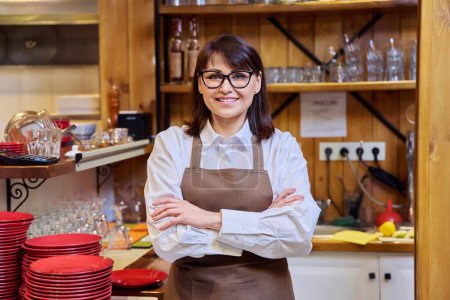Photo for Portrait of middle aged woman bistro restaurant manager owner in an apron, confident smiling looking at camera with crossed arms. Small business, service, work, staff concept - Royalty Free Image