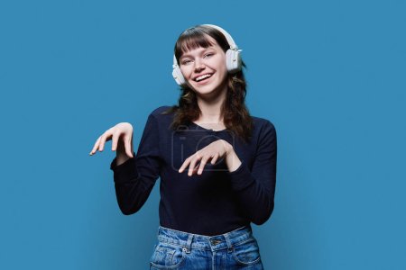 Photo for Cheerful attractive young female in headphones dancing trendy dance, on blue color studio background. Modern youth, fashion, music, dancing, fun, lifestyle, people concept - Royalty Free Image