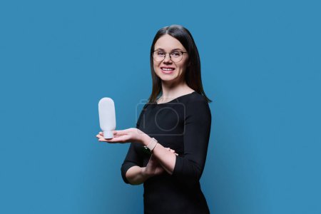 Young woman holding white clean tube of body face cream, on blue studio background. Advertising sale of cosmetics for care, treatment, dermatology, cosmetology, beauty industry concept