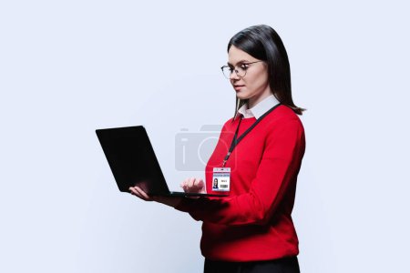 Joven maestra mentora con placa de identificación del empleado del centro educativo, en rojo, usando computadora portátil sobre fondo blanco. Concepto de enseñanza de formación de trabajo organizacional administrativo