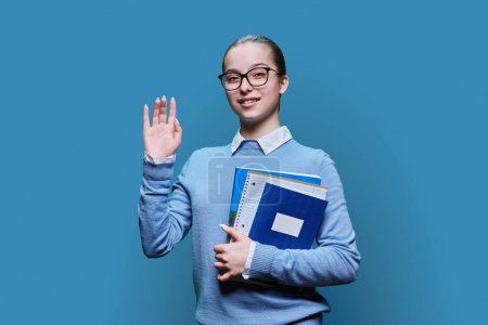 Téléchargez les photos : Portrait d'une lycéenne souriante avec des cahiers de notes dans les mains sur fond de studio bleu. Smart amical adolescent femme agitant à la caméra. Adolescence, éducation, concept d'étude - en image libre de droit