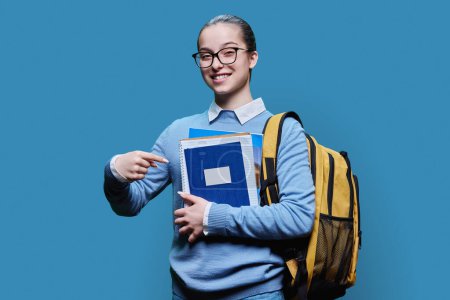 Téléchargez les photos : Portrait d'une adolescente lycéenne sur fond de studio bleu. Femme souriante en chandail avec sac à dos regardant la caméra. Éducation, adolescence, biens et services pour adolescents - en image libre de droit