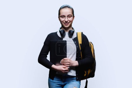 Téléchargez les photos : Étudiante adolescente avec sac à dos tenant un ordinateur portable sur fond de studio blanc. Jeune femme souriante dans des lunettes avec écouteurs regardant la caméra. École secondaire, adolescence, concept d'éducation - en image libre de droit