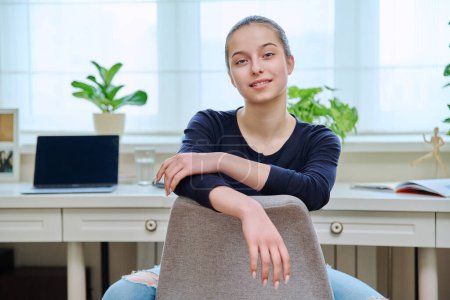 Téléchargez les photos : Portrait d'une adolescente souriante assise sur une chaise intérieur de la maison. Adolescente 15, 16 ans regardant à la caméra, fenêtre de bureau avec fond d'ordinateur portable. Adolescence style de vie lycéens - en image libre de droit