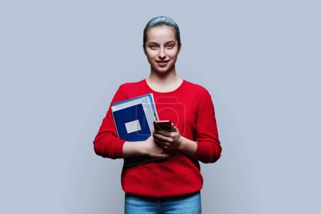 Téléchargez les photos : Portrait d'une adolescente lycéenne sur fond gris. Femme souriante avec smartphone tenant des manuels regardant la caméra. Éducation adolescence applications mobiles pour l'étude, les services les adolescents - en image libre de droit