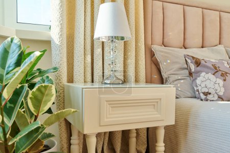 Photo for Bedside table, lamp with lampshade, in the interior of a classic bedroom in light pink colors, in warm sunlight. Comfort, home, style, decor, design concept - Royalty Free Image