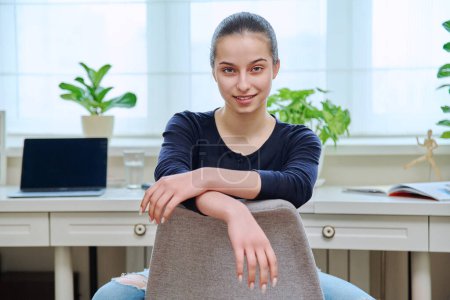 Téléchargez les photos : Portrait d'une adolescente souriante assise sur une chaise intérieur de la maison. Adolescente 15, 16 ans regardant à la caméra, fenêtre de bureau avec fond d'ordinateur portable. Adolescence style de vie lycéens - en image libre de droit