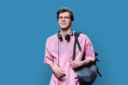 Téléchargez les photos : Jeune hipster masculin tendance en t-shirt rose sur fond studio bleu. Beau gars en tricot lunettes chapeau avec casque sac à dos regardant la caméra. Mode, mode de vie, jeune 18-20 ans - en image libre de droit