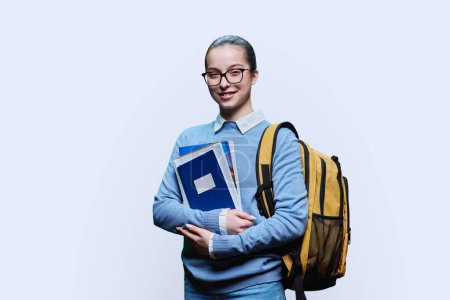 Téléchargez les photos : Portrait d'une adolescente lycéenne sur fond de studio blanc. Femme souriante en chandail bleu avec sac à dos regardant la caméra. Éducation, adolescence, biens et services pour adolescents - en image libre de droit