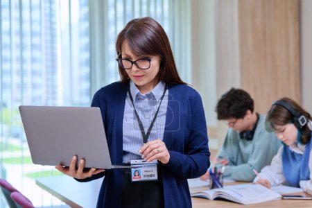 Photo for Portrait of confident middle aged female teacher tutor schoolmaster educator holding laptop looking at screen, in college classroom with students at desk. Education learning language courses knowledge - Royalty Free Image