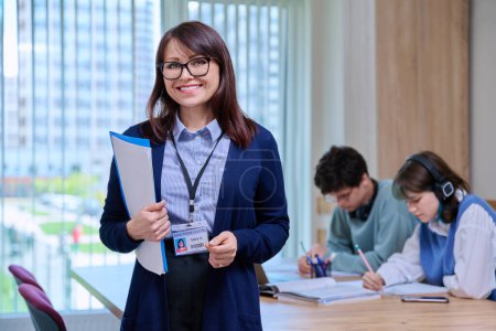 Photo for Portrait of confident middle aged female teacher schoolmaster tutor educator looking at camera, in college classroom with students at desk. Education, learning, language courses, knowledge concept - Royalty Free Image