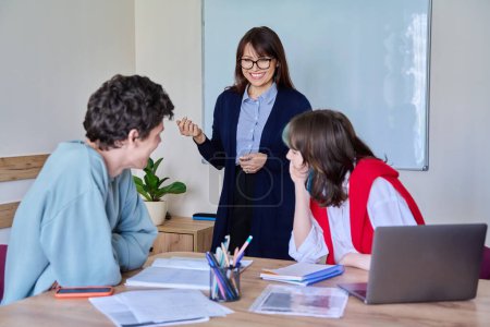 Photo for Middle aged female teacher giving lesson to teenage college students sitting at table. Education, knowledge, learning, studying, language courses, youth concept - Royalty Free Image