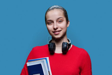 Téléchargez les photos : Portrait d'une lycéenne souriante avec des cahiers de notes dans les mains sur fond de studio bleu. Adolescente intelligente et amicale en rouge regardant la caméra. Adolescence, éducation, concept d'étude - en image libre de droit