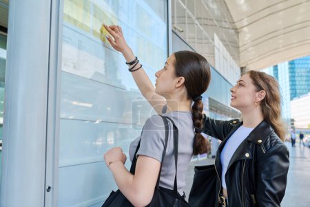 Photo for Two young female students reading transport timetable on scoreboard. Public transport, passengers, urban lifestyle concept - Royalty Free Image