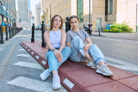 Téléchargez les photos : Deux adolescentes souriantes regardant la caméra, le style urbain moderne, le fond des bâtiments. Filles à la mode assis sur le trottoir près de la route. Jeunesse, mode, style, ville, concept de style de vie - en image libre de droit