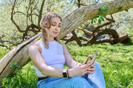 Young female sitting on the grass in the park, relaxing with smartphone. Mobile applications for leisure, learning, shopping, relaxation, online internet technology