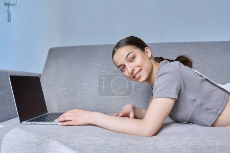 Téléchargez les photos : Adolescente à la maison sur le canapé en utilisant un ordinateur portable, souriant en regardant la caméra. Technologie pour apprendre la communication de loisirs, mode de vie moderne, concept d'adolescence - en image libre de droit
