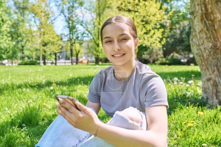 Téléchargez les photos : Jeune femme assise sur l'herbe dans le parc, se relaxant avec un smartphone, souriant en regardant la caméra. Applications mobiles pour les loisirs, l'apprentissage, le shopping, la relaxation, la technologie Internet en ligne - en image libre de droit