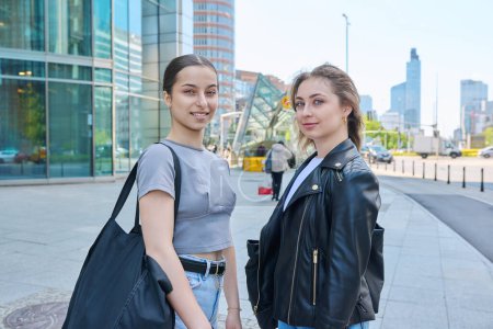 Téléchargez les photos : Deux adolescentes souriantes regardant la caméra, arrière-plan urbain moderne. Jeunesse, ville, mode de vie, filles âge collégial - en image libre de droit