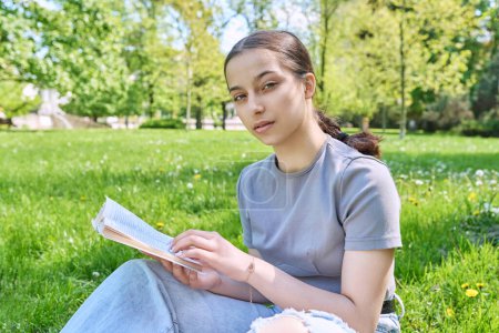 Téléchargez les photos : Adolescente lycéenne livre de lecture assis sur l'herbe dans le parc. Souriant jeune femme regardant la caméra - en image libre de droit