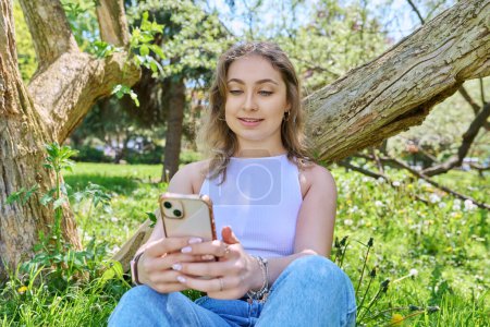 Young female sitting on the grass in the park, relaxing with smartphone. Mobile applications for leisure, learning, shopping, relaxation, online internet technology