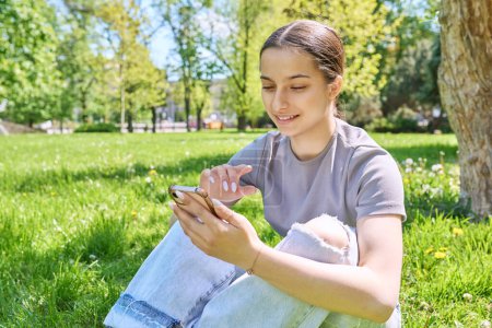 Téléchargez les photos : Jeune femme assise sur l'herbe dans le parc, se détendre avec smartphone. Applications mobiles pour les loisirs, l'apprentissage, le shopping, la relaxation, la technologie Internet en ligne - en image libre de droit