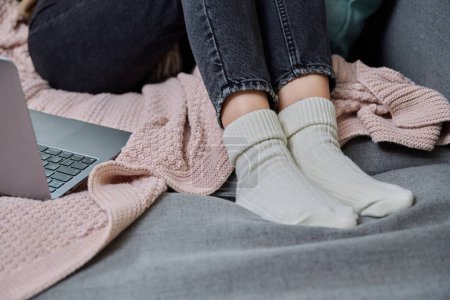 Close-up of legs in cozy woolen socks, laptop plaid sofa. Cold autumn winter season, holidays, relax, comfort, coziness, lifestyle, seasonality concept