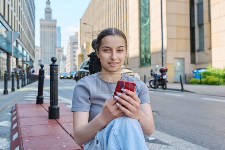 Téléchargez les photos : Jeune adolescente souriante avec smartphone regardant le téléphone dans la rue de la ville européenne moderne. École secondaire adolescence mode style de vie des jeunes, applications mobiles technologie Internet en ligne - en image libre de droit