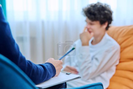 Photo for Close-up of female psychologists hands with clipboard making notes, in individual therapy session. Young male patient in chair out of focus. Psychology psychotherapy treatment mental health concept - Royalty Free Image