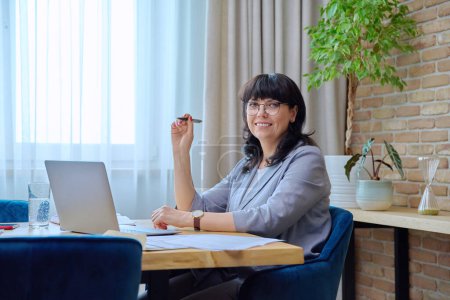 Photo for Working confident smiling business mature woman sitting at desk using laptop computer business papers contracts, looking at camera in office. 50s female leader manager financial director workplace - Royalty Free Image