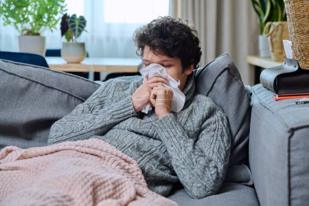 Téléchargez les photos : Jeune homme malade éternuant dans les tissus nasaux, ayant des signes de maladie infectieuse respiratoire, en pull en laine assis sur le canapé à la maison. nez qui coule, toux, saison froide - en image libre de droit