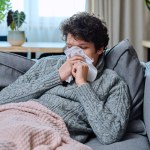 Sick young guy sneezing into nasal tissue, having signs of respiratory infectious disease, in woolen sweater sitting on couch at home. Runny nose, cough, cold season