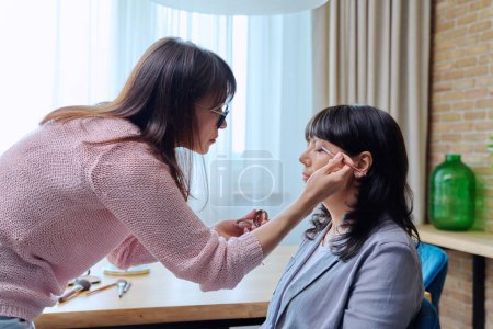 Téléchargez les photos : Maquilleuse faisant du maquillage pour une femme d'affaires mature, peint les yeux avec un pinceau avec des ombres. Beauté, cosmétiques, soins, visage, femme de 50 ans - en image libre de droit